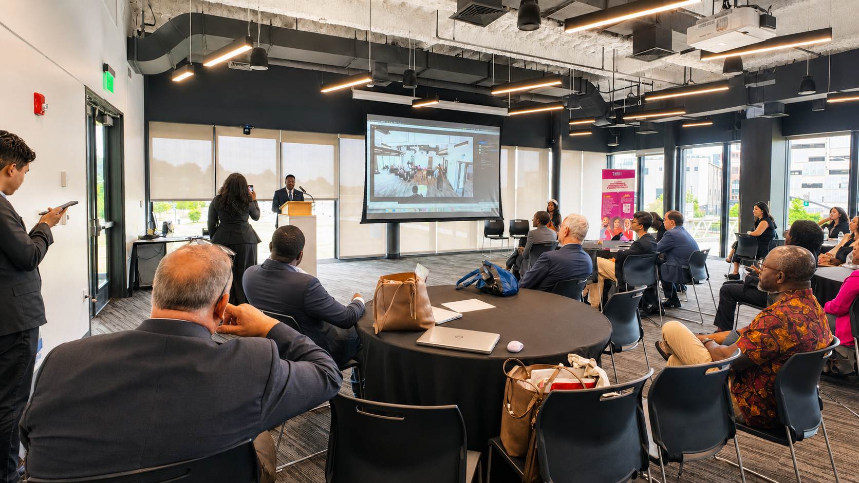 A speaker at a podium addresses attendees seated at round tables during a GHPAC business event, with remote participants shown on a large projection screen